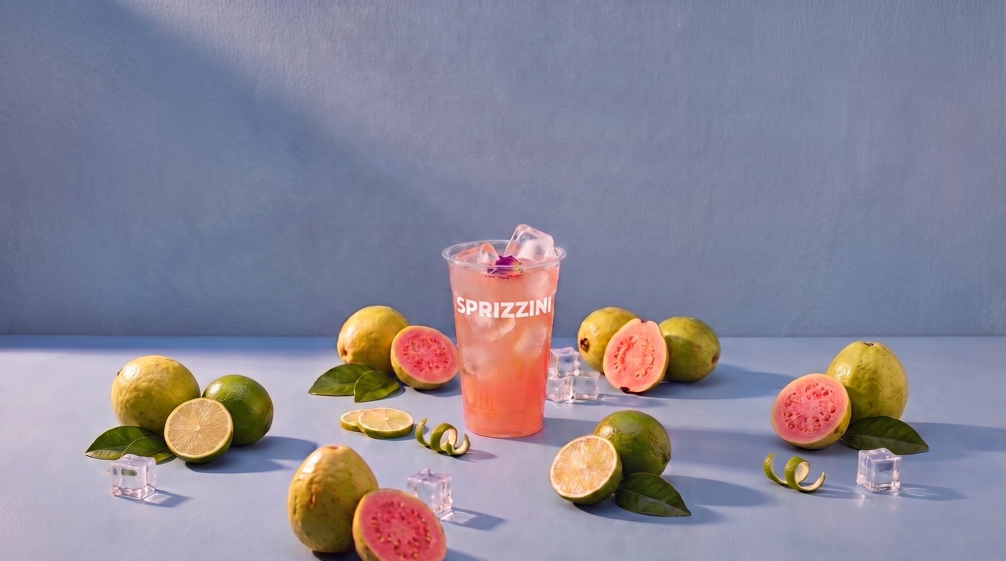 A Guava Rose in a branded Sprizzini cup on a dusty blue studio set, guavas and limes around it